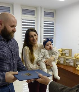 Burmistrz Janusz Reszelewski wręcza rodzicom z niemowlęciem pamiątkowy dyplom z gratulacjami. Dyplom odbiera tata. Mama trzyma dziecko. Na blacie stoją pudełka i pluszowe misie.