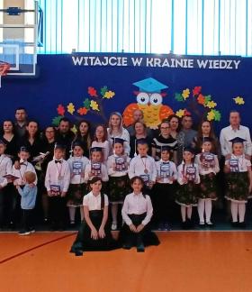 Grupowo pozujące dzieci w galowych strojach, nauczyciele i rodzice podczas uroczystości szkolnej w sali gimnastycznej SP w Brzustowcu. W tle niebieska ściana z napisem „WITAJCIE W KRAINIE WIEDZY” oraz dekoracją z sową i jesiennymi liśćmi.