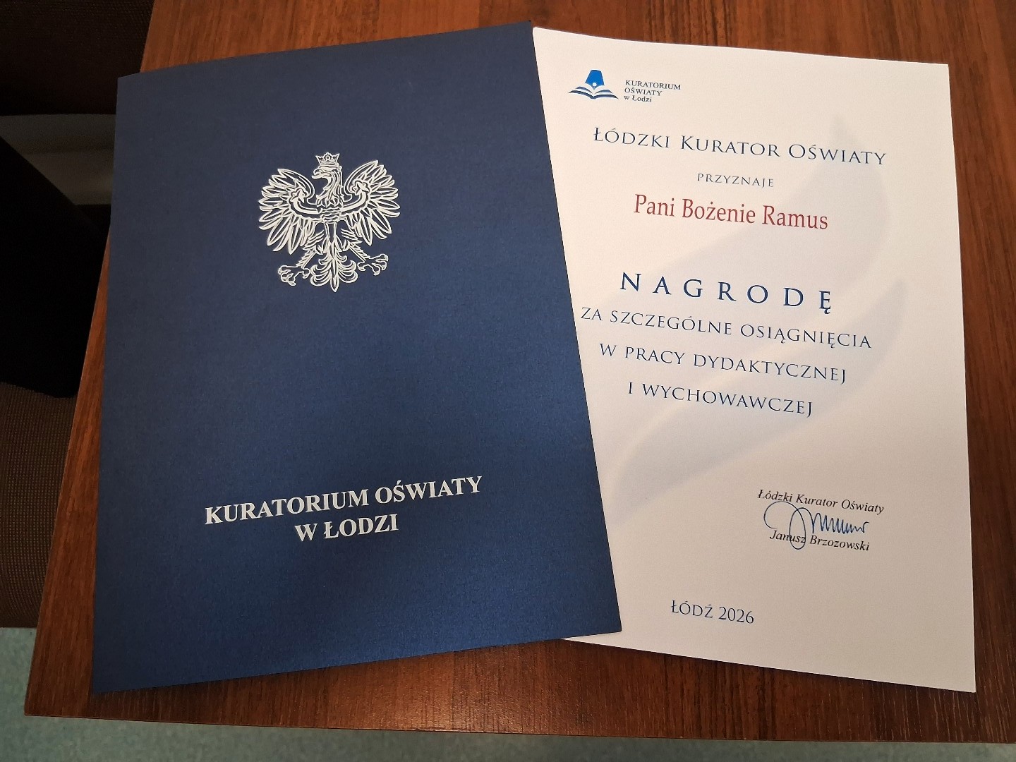 Dyplom nagrody Łódzkiego Kuratora Oświaty dla Pani Bożeny Ramus. Ciemnoniebieska okładka z białym godłem Polski i napisem „KURATORIUM OŚWIATY W ŁODZI” spoczywa obok dyplomu na drewnianym blacie.