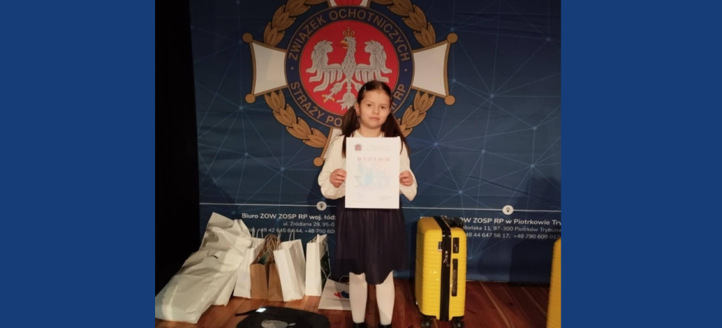 Trzymająca dyplom dziewczynka stoi na tle loga Związku OSP RP. Po obydwu stronach dziecka nagrody rzeczowe