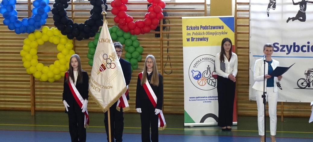 Oficjalne otwarcie gali w hali sportowej. Od lewej: trzyosobowy szkolny poczet sztandarowy na tle kół olimpijskich, uczennica Michalina Jedynak i Dyrektor SP w Drzewicy Agnieszka Bogatek