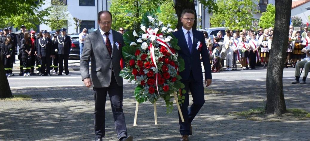 Dwaj pracownicy Urzędu Miejskiego w Drzewicy zmierzają pod Pomnik Pamięci, niosąc wieniec z białymi i czerwonymi kwiatami