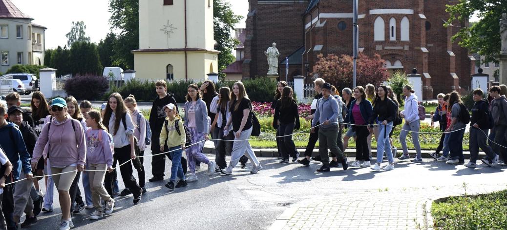 Grupa pielgrzymów wychodzi na trasę. Maszeruje ulicą na placu Wolności w Drzewicy. W tle m.in. kościół i dzwonnica.