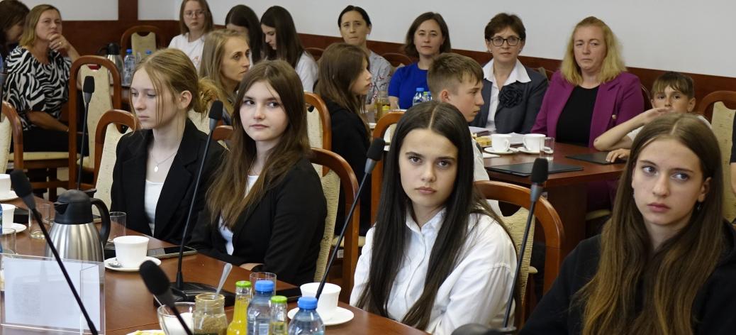 Uczestnicy sesji siedzą przy stołach w sali konferencyjnej Urzędu Miejskiego w Drzewicy. W centrum zdjęcia młodzieżowe radne, z tyłu m.in. nauczycielki.