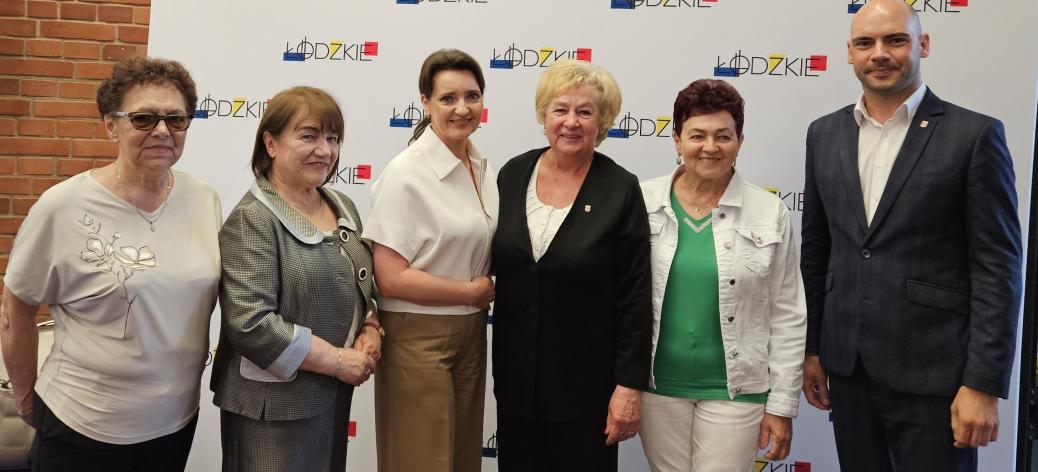 Przed ścianką promocyjną Urzędu Marszałkowskiego Województwa Łódzkiego stoją od lewej: radna senioralna Zdzisława Majchrowska, Przewodnicząca Rady Seniorów Województwa Łódzkiego Krystyna Ozga, Minister ds. Polityki Senioralnej Marzena Okła-Drewnowicz, Przewodnicząca Miejskiej Rady Seniorów w Drzewicy Barbara Oliwa, radna senioralna Jadwiga Szcześniak, Zastępca Burmistrza Drzewicy Dominik Niemirski