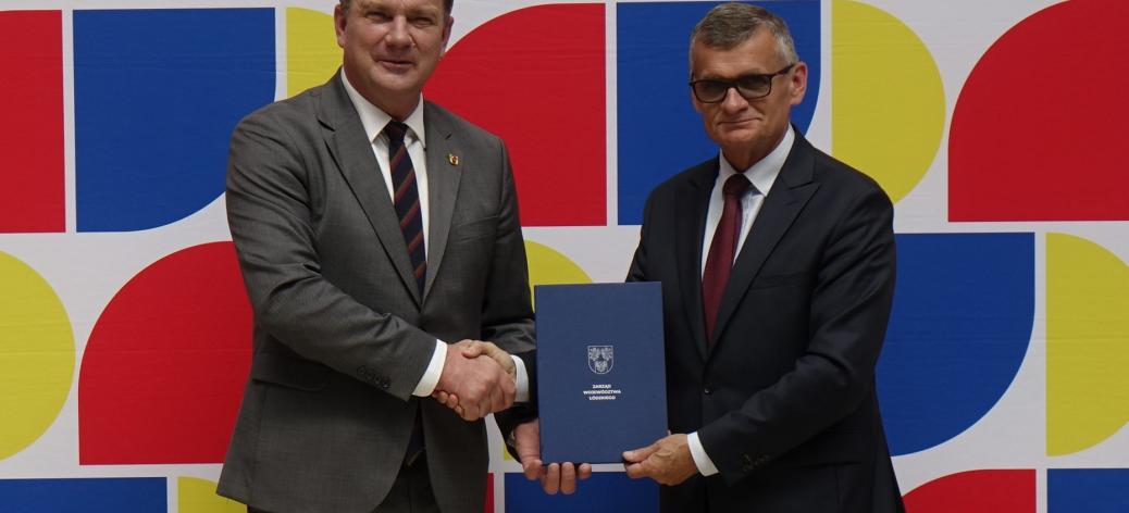 Na tle ścianki promocyjnej Województwa Łódzkiego w uścisku dłoni Członek Zarządu Województwa Łódzkiego Artur Bagieński oraz Burmistrz Drzewicy Janusz Reszelewski