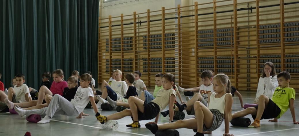 Na zdjęciu uczniowie trzecich klas szkół podstawowych z terenu Gminy Drzewica znajdują się w hali sportowej SP w Drzewicy przy ul. Staszica. Wykonują ćwiczenia praktyczne z rollerami. Rolują łydkę.