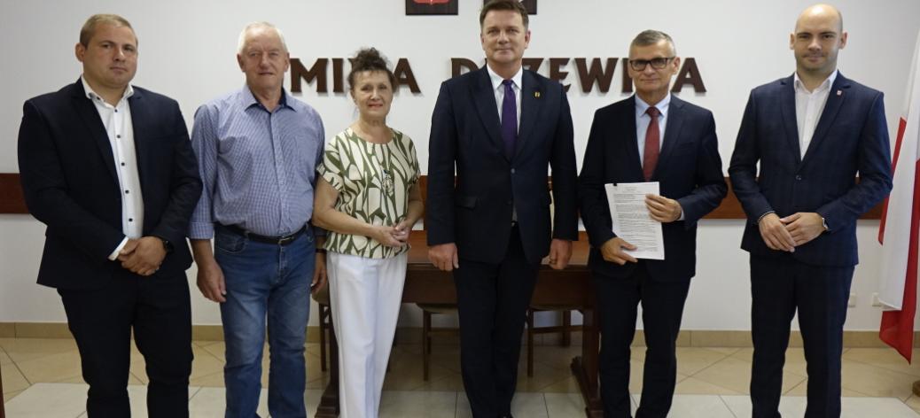 3.	Wspólne zdjęcie. W sali konferencyjnej Urzędu Miejskiego w Drzewicy, na tle ściany z napisem Gmina Drzewica, herbem Drzewicy oraz godłem Rzeczpospolitej Polskiej stoją: (od lewej): Sołtys Miejscowości Świerczyna Karol Siedlecki, Sołtys Miejscowości Idzikowice Roman Mastalerek, Skarbnik Gminy Drzewica Irena Jóźwicka, Wicemarszałek Województwa Łódzkiego Artur Bagieński, Burmistrz Drzewicy Janusz Reszelewski, Zastępca Burmistrza Drzewicy Dominik Niemirski. 