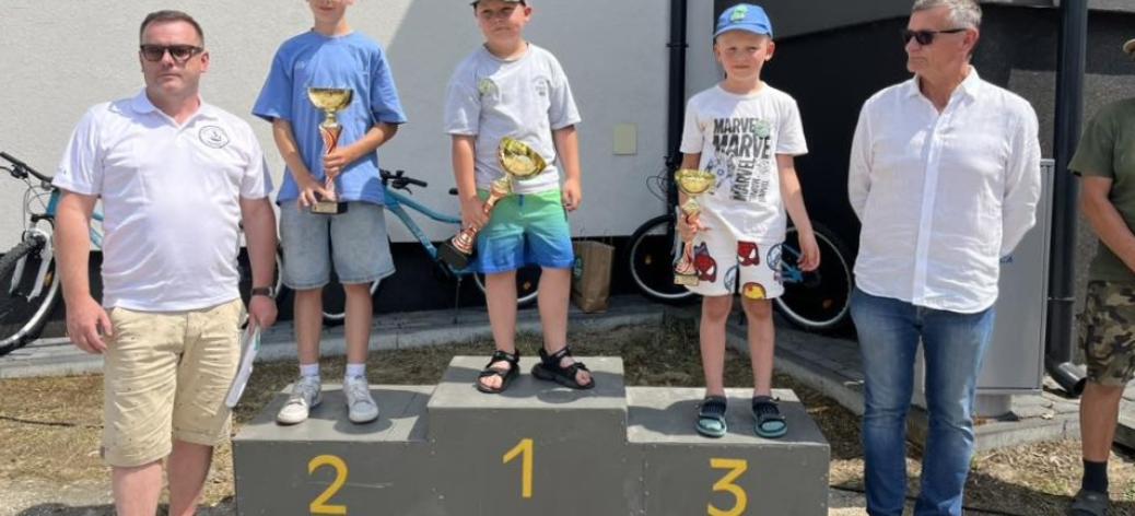 Dekoracja w kategorii chłopców. Na podium trzej młodzi wędkarze z pucharami. Obok po lewej Prezes Koła nr 3 PZW w Drzewicy Ziemowit Jaciubek, po prawej Burmistrz Drzewicy Janusz Reszelewski