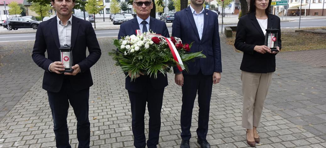 Gminna delegacja w parku na placu Wolności. Od lewej stoją: Sekretarz Miasta Andrzej Krzyżanowski ze zniczem, Burmistrz Drzewicy Janusz Reszelewski z wiązanką kwiatów, Zastępca Burmistrza Dominik Niemirski, Przewodnicząca Rady Miejskiej w Drzewicy Dorota Wiktorowicz ze zniczem.