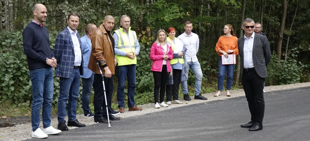 Na wyremontowanej drodze powiatowej stoją uczestnicy odbioru inwestycji – 11 osób. W tle las