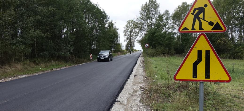 Widok remontowanej drogi powiatowej z nową warstwą ścieralną. Po drodze jedzie auto. Z prawej znaki informujące o robotach drogowych