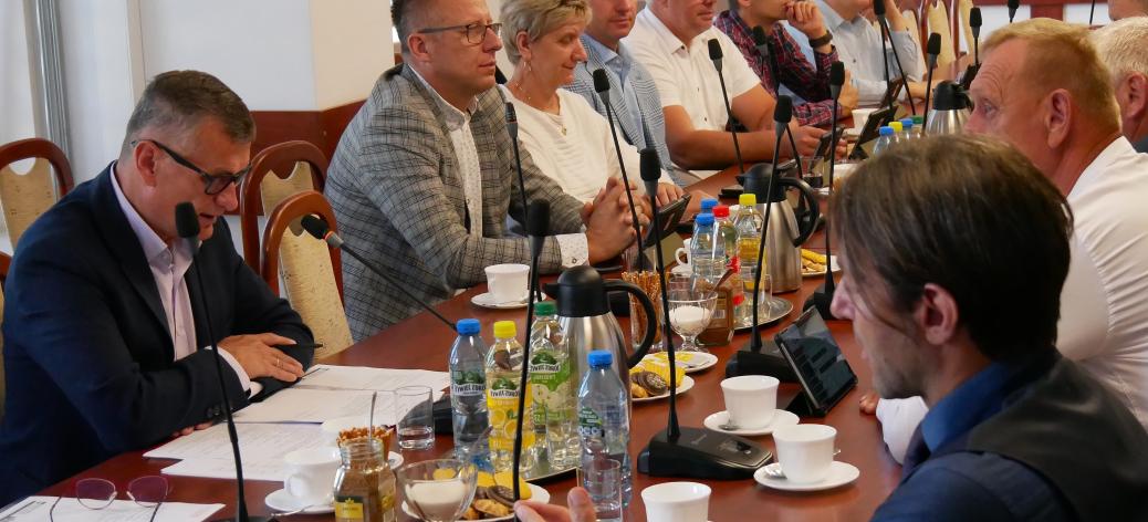 Przy stole w sali konferencyjnej UM w Drzewicy siedzą uczestnicy sesji. Głos zabiera Burmistrz Drzewicy Janusz Reszelewski. Słuchają radni i Sekretarz Miasta