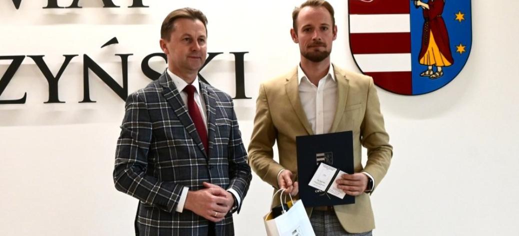 Od lewej stoją Starosta Opoczyński Marcin Baranowski i Piotr Bajerowski, który trzyma dokmenty i upominek. W tle ściana z herbem i częścią nazwy powiatu opoczyńskiego