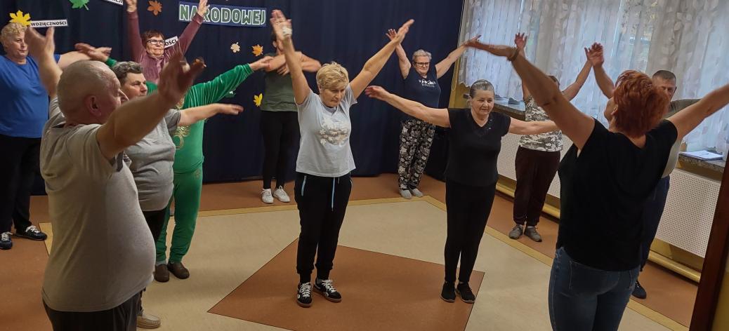 Grupa seniorów na zajęciach fitness w SP w Idzikowicach. Na pierwszym planie instruktor zajęć Alina Szymańska, na planie dalszym uczestnicy zajęć patrzący na instruktora wykonują ćwiczenia