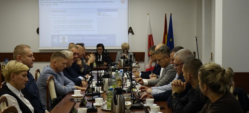 Obrady Nadzwyczajnej Sesji Rady Miejskiej w Drzewicy na sali konferencyjnej Urzędu Miejskiego w Drzewicy. Radni na pierwszym planie zasiadają przy stole. Na drugim planie prezydium rady przy stole.  