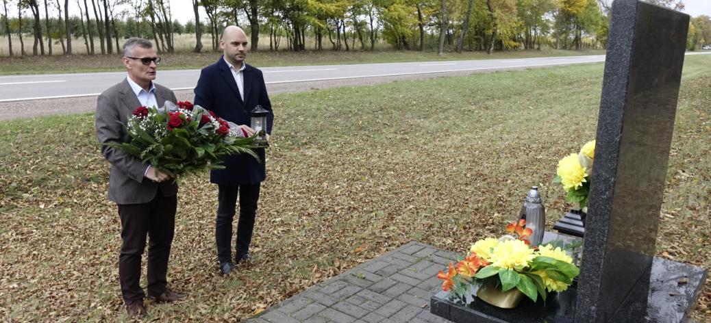 Pomnik nagrobny, po lewej stoją Burmistrz Drzewicy Janusz Reszelewski trzymający wieniec i Zastępca Burmistrza Drzewicy Dominik Niemirski trzymający znicz. W tle pas drogi wojewódzkiej. 