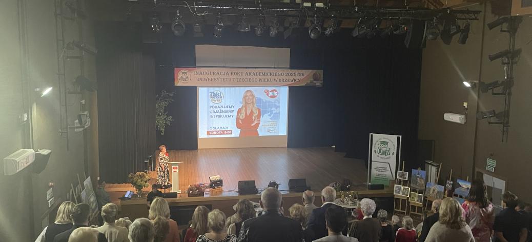 Inauguracja roku akademickiego UTW w Drzewicy. Zdjęcie z góry publiczności. Zgromadzeni stoją podczas odśpiewania hymnu „Gaudeamus igitur”. W tle scena.