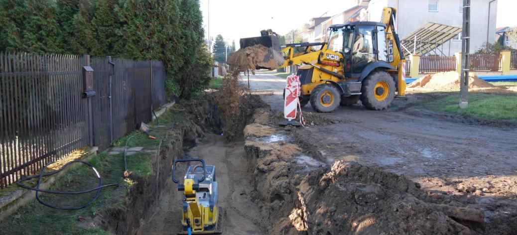 Przebudowa sieci wodociągowej na Os. Słonecznym w Drzewicy. Na zdjęciu wykop, w środku którego znajduje się zagęszczarka. W tle koparko-ładowarka zasypuje wykop drobnym piaskiem. Wokoło zabudowania przy ulicy. 