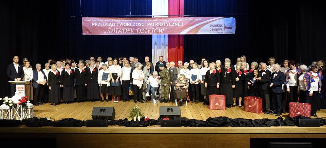  Wspólne zdjęcie wykonawców i organizatorów przeglądu po uroczystej dekoracji. Na scenie stoi kilkadziesiąt osób, nad którymi wisi baner z nazwą imprezy