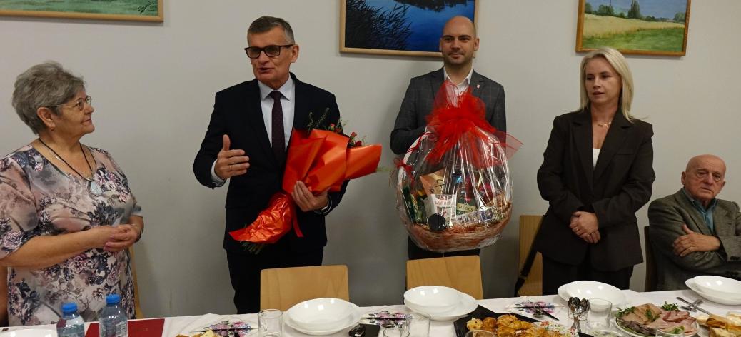 Podczas oficjalnej części uroczystości przemawia trzymający bukiet kwiatów Burmistrz Drzewicy Janusz Reszelewski. Po lewej stronie Burmistrza stoi Przewodnicząca Koła Emerytów i Rencistów Jadwiga Gapys, po prawej stoją Zastępca Burmistrza Dominik Niemirski z koszem słodkości i Prezes CAL w Drzewicy Edyta Bartos, a także siedzi jeden z emerytów.  