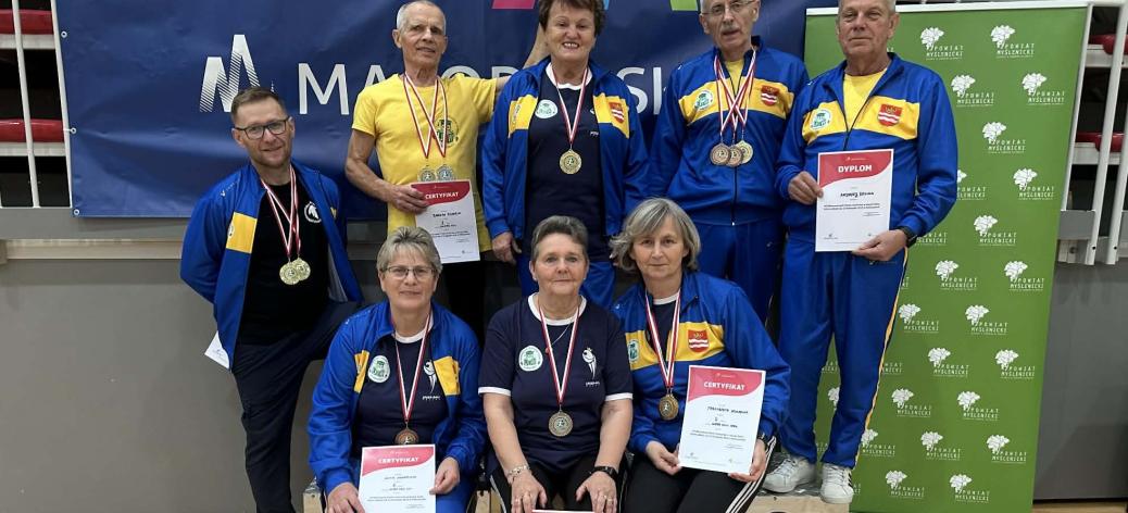 Na podium stoją i siedzą ubrani w żółto-niebieskie dresy reprezentanci UTW w Drzewicy - osiem osób z medalami i dyplomami