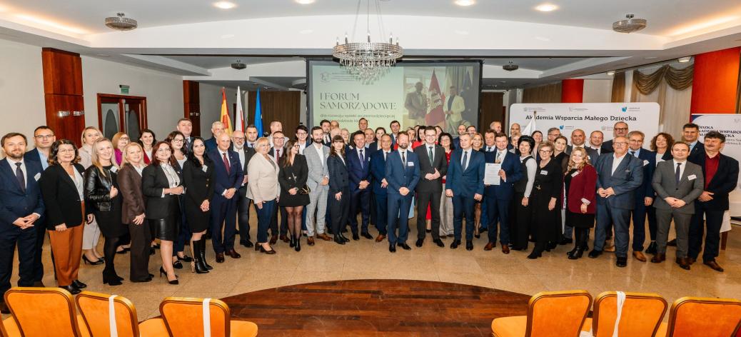 Wspólne zdjęcie kilkudziesięcioosobowowej grupy uczestników Forum stojących w przestronnej sali konferencyjnej. W tle ekran, flagi, rull-upy, na górze żyrandol