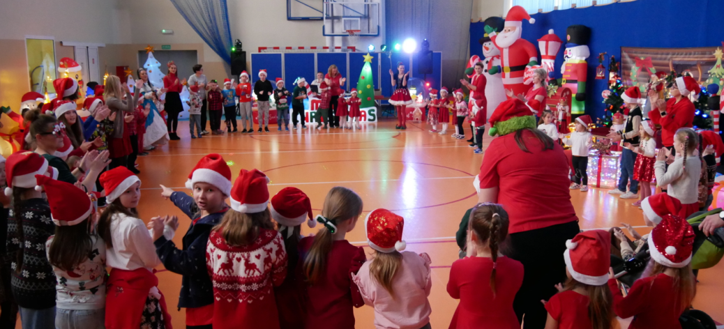 Mikołajki w SP w Brzustowcu. Grupa uczniów wraz z pracownikami szkoły zgromadzona w okręgu w sali gimnastycznej. Wokoło dmuchane dekoracje świąteczne – bałwany, mikołaje, renifery, żołnierzyki.