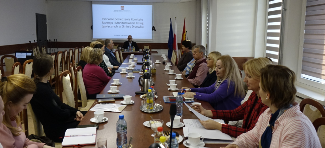 Spotkanie komitetu w Sali konferencyjnej Urzędu Miejskiego w Drzewicy. Na pierwszym planie przy stole głównym członkowie komitetu. W tle przy stole prezydialnym Zastępca Burmistrza Drzewicy Dominik Niemirski. Na ścianie za jego plecami na ekranie prezentacja multimedialna. 