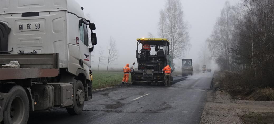 Trwają prace związane z modernizacją drogi powiatowej Krzczonów-Jelnia. Na drodze stoją: na pierwszym planie samochód ciężarowy, dalej rozściełacz do asfaltu a przy nim pracujący drogowcy. W tle dwa walce na asfalcie. 