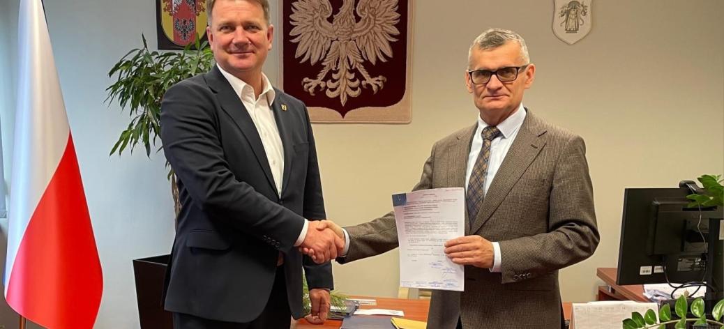 Członek Zarządu Woj. Łódzkiego Artur Bagieński i Burmistrz Drzewicy Janusz Reszelewski podają sobie dłonie. Burmistrz trzyma aneks. Po lewej flaga Polski, w tle godła Polski i Woj. Łódzkiego