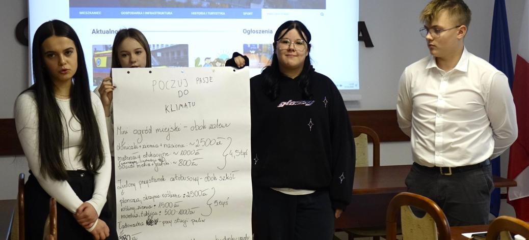 W sali konferencyjnej UM na tle ekranu stoi czworo uczniów. Jedna z dziewcząt trzyma zapisany duży arkusz papieru