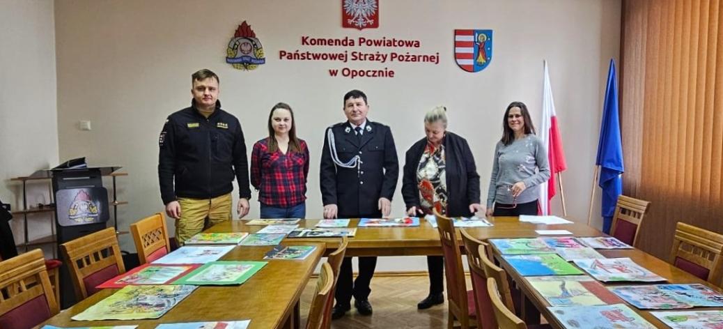 W sali konferencyjnej KP PSP w Opocznie za stołami, na których rozłożone są prace plastyczne, stoi pięć osób - dwóch strażaków i trzy panie.  W środku Gminny Komendant ZOSP RP w Drzewicy Marian Kalużny
