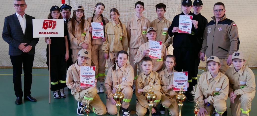 Młodzieżowe Drużyny Pożarnicze z OSP Domaszno wraz z opiekunami prezentują zdobyte trofea. Po lewej stoi Burmistrz Drzewicy.  W tle bramka do piłki ręcznej.