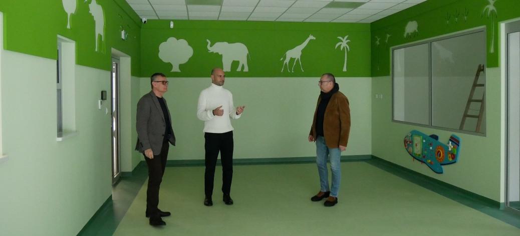 Pomalowane w odcieniach zieleni pomieszczenie bawialni oglądają od lewej: Burmistrz Drzewicy Janusz Reszelewski, Zastępca Burmistrza Dominik Niemirski oraz Kierownik Referatu Gospodarki Komunalnej, Budownictwa i Inwestycji Urzędu Miejskiego w Drzewicy