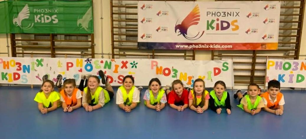 W hali sportowej dziesięcioro dzieci leży na parkiecie przodem do fotografa. W tle drabinki i banery Pho3nix Kids.