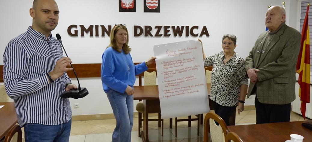 W sali konferencyjnej UM na tle napisu Gmina Drzewica stoi mówiący do mikrofonu Zastępca Burmistrza Drzewicy oraz troje radnych Miejskiej Rady Seniorów. Dwie radne trzymają zapisany arkusz papieru