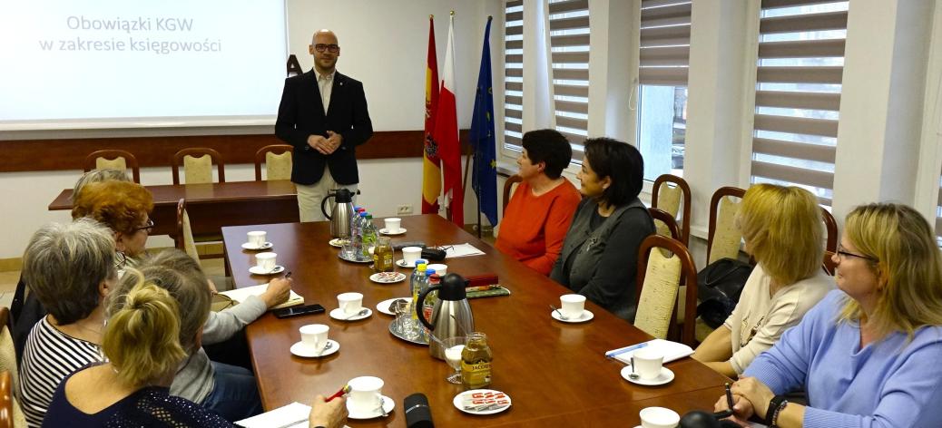  Zastępca Burmistrza Drzewicy Dominik Niemirski prowadzi szkolenie dla pań z KGW siedzących przy stole konferencyjnym. W  tle ekran z napisem ,,O obowiązkach KGW w księgowości'' oraz flagi