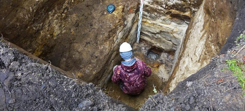 W głębokim wykopie, kucając pracuje archeolog z kaskiem na głowie (widok na jego plecy). Powyżej miarka głębokości.