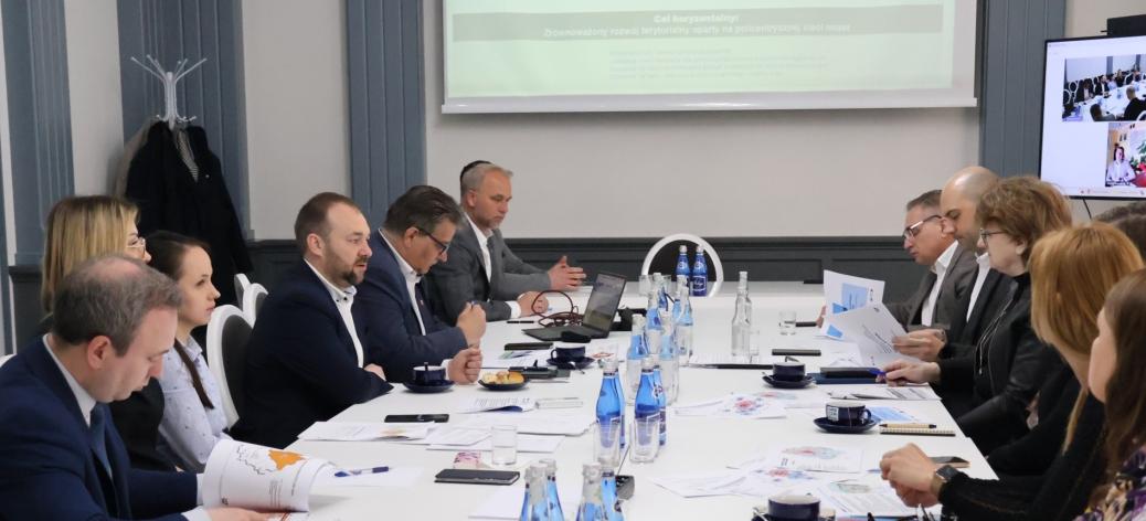 Na zdjęciu spotkanie odbywające się w sali konferencyjnej. Przy długim stole siedzi kilkanaście osób, przeglądają dokumenty, notują oraz słuchają osoby mówiącej.  W tle widać ekran projekcyjny z wyświetloną prezentacją.