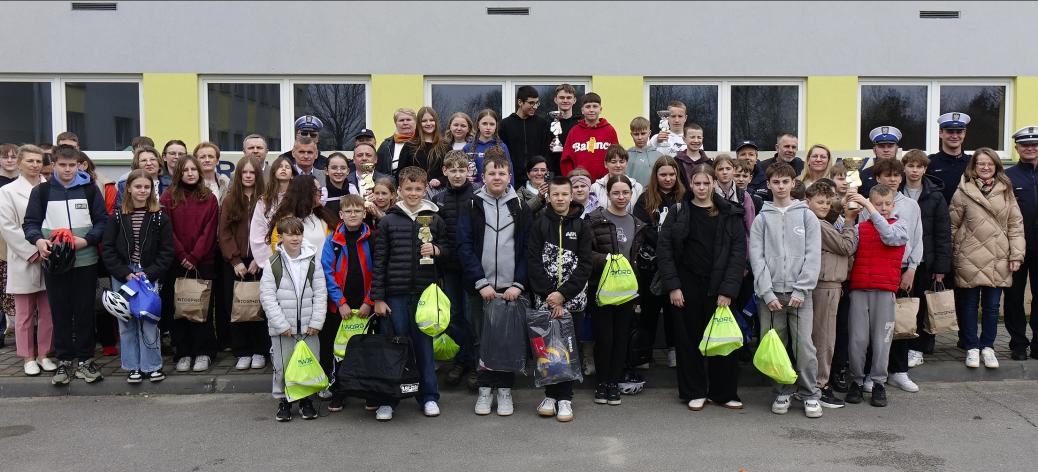 Duża grupa dzieci, młodzieży oraz dorosłych ustawiona w kilku rzędach przed budynkiem o jasnej elewacji z zielonymi elementami. Uczestnicy stoją blisko siebie i pozują do wspólnego zdjęcia. Wśród nich widoczni są uczniowie oraz opiekunowie, a także kilku umundurowanych policjantów