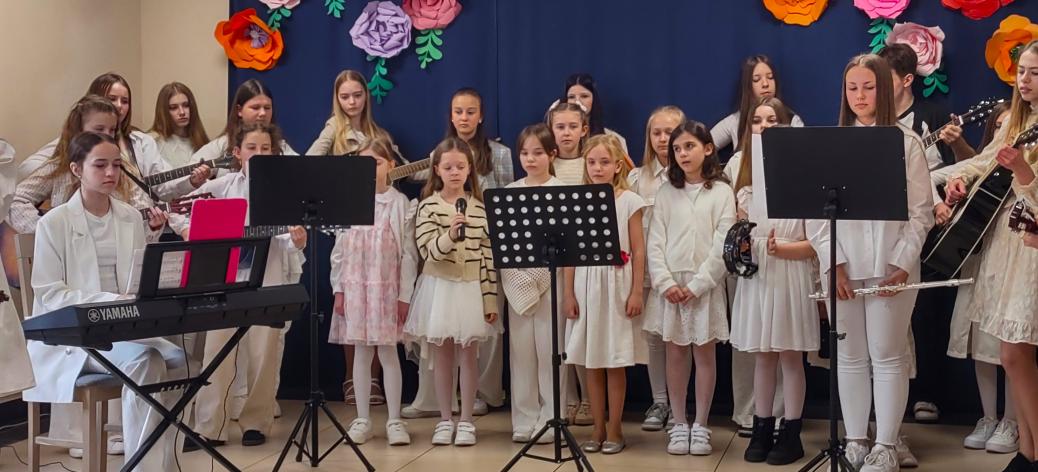 Grupa dzieci i młodzieży stoi na niewielkiej scenie podczas występu muzycznego w siedzibie PDPS w Drzewicy. Uczestnicy ubrani są głównie na biało. Na pierwszym planie kilka dziewczynek śpiewa do mikrofonu ustawionego na statywie. Obok znajdują się pulpity na nuty. Po lewej stronie osoba siedzi przy keyboardzie. W tle część uczestników gra na gitarach. Za nimi widoczna jest granatowa kurtyna ozdobiona dużymi, kolorowymi, papierowymi kwiatami.