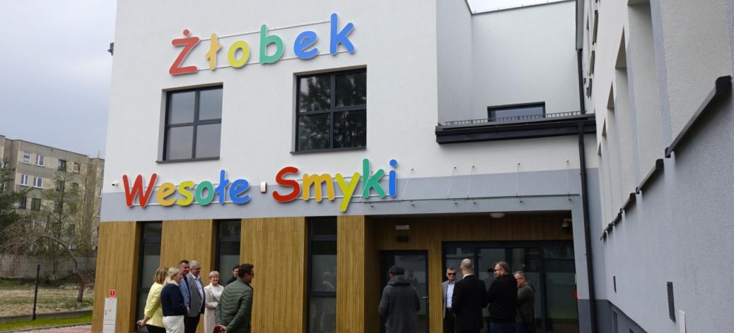 Grupa osób w strojach wizytowych i codziennych stoi przed nowoczesnym, białym budynkiem żłobka „Wesołe Smyki”. Budynek ma kolorowy napis nad wejściem i drewnopodobne panele na parterze.