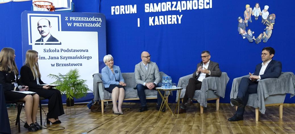 Na zdjęciu widoczny jest panel dyskusyjny podczas Forum Samorządności i Kariery w Szkole Podstawowej im. Jana Szymańskiego w Brzustowcu. Na sofie, fotelach bądź krzesłach siedzi sześć osób – czworo panelistów oraz dwie moderatorki po lewej stronie. Burmistrz Drzewicy trzyma mikrofon i zabiera głos. W tle znajduje się niebieska ścianka z napisem „Forum Samorządności i Kariery” oraz grafika przedstawiająca różne zawody. Po lewej stronie widoczny jest baner szkoły z portretem patrona.