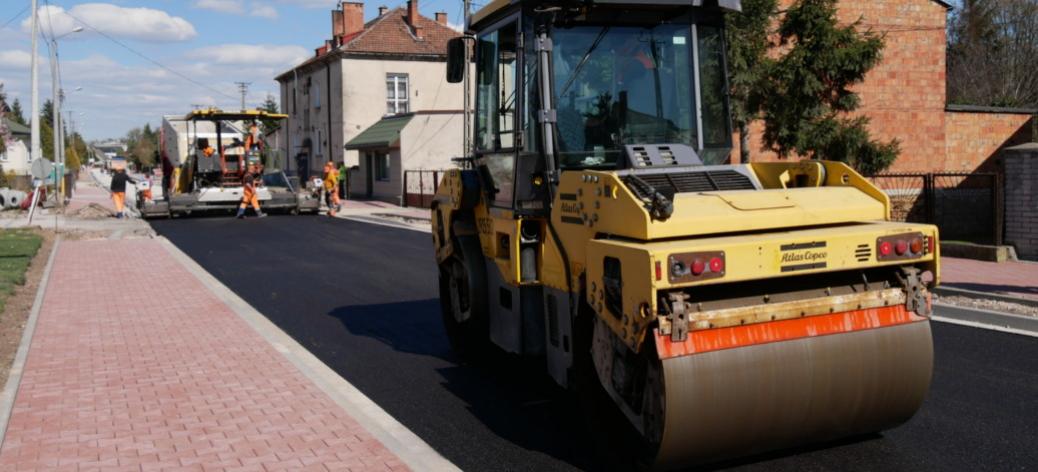 Modernizacja drogi z układaniem nowej nawierzchni asfaltowej przy użyciu specjalistycznych maszyn. Na zdjęciu widoczny walec drogowy oraz pracownicy w odzieży ostrzegawczej podczas prac budowlanych.