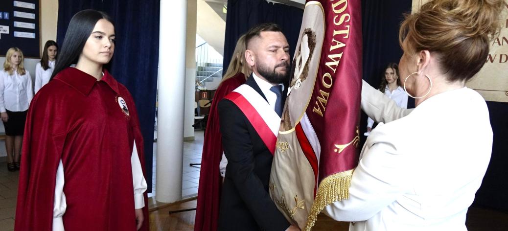 Wnętrze szkoły podczas uroczystości. Mężczyzna z biało-czerwoną szarfą trzyma sztandar szkoły w kolorze czerwonym z haftem. Naprzeciw stoi kobieta w jasnym stroju, poprawiająca lub przekazująca sztandar. Obok  uczennica w czerwonej pelerynie, patrząca w stronę sztandaru. 