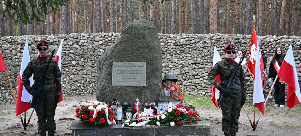 Uroczystość patriotyczna w lesie. Dwie żołnierki w mundurach polowych i bordowych beretach trzymają wartę z bronią po obu stronach pomnika-głazu z pamiątkową tablicą. U podnóża monumentu leżą liczne wieńce i znicze, a obok widoczne są polskie flagi. W tle znajduje się kamienny mur i wysoki las sosnowy.