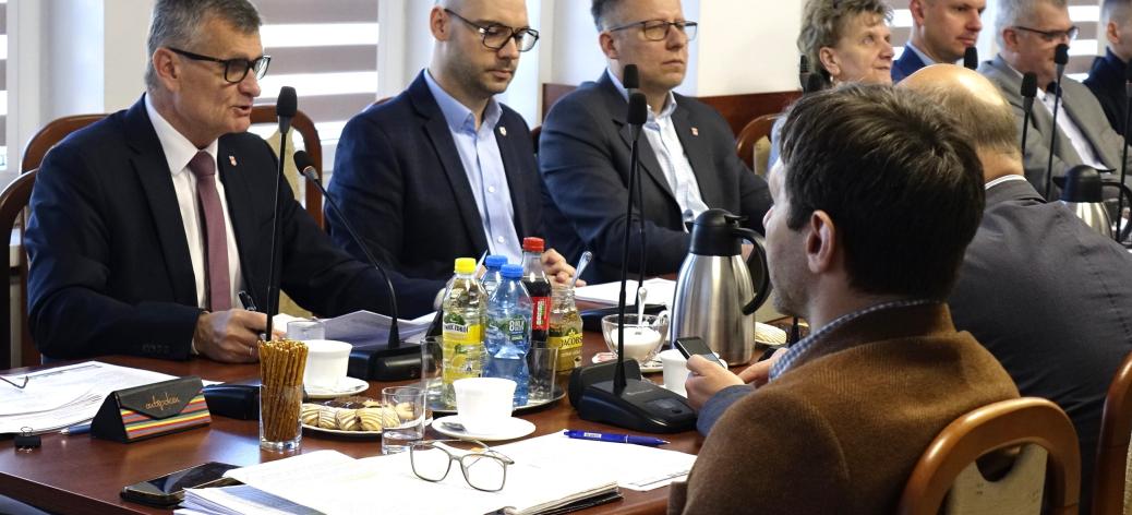 Przy stole z mikrofonami i dokumentami siedzi kilka osób w formalnych strojach – Burmistrz i Zastępca Burmistrza Drzewicy oraz radni. Głos zabiera Burmistrz Drzewicy, pozostali słuchają. Na stole widoczne są napoje i materiały do pracy.