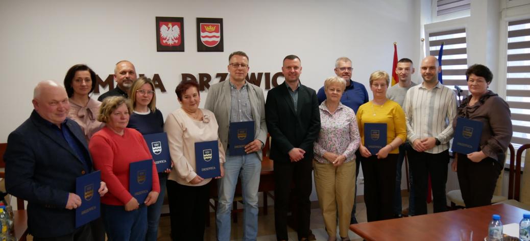 Zdjęcie grupowe przedstawicieli kół gospodyń oraz władz samorządowych w sali konferencyjnej Urzędu Miejskiego w Drzewicy. Uczestnicy trzymają pamiątkowe dyplomy w niebieskich okładkach z herbem gminy.