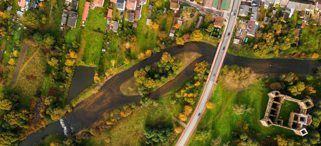 Widok z lotu ptaka na jesienny krajobraz w Drzewicy, przedstawiający ruiny gotycko-renesansowego zamku, rzekę Drzewiczkę z jazem, które otoczone są kolorowymi drzewami i zabudową miejską.